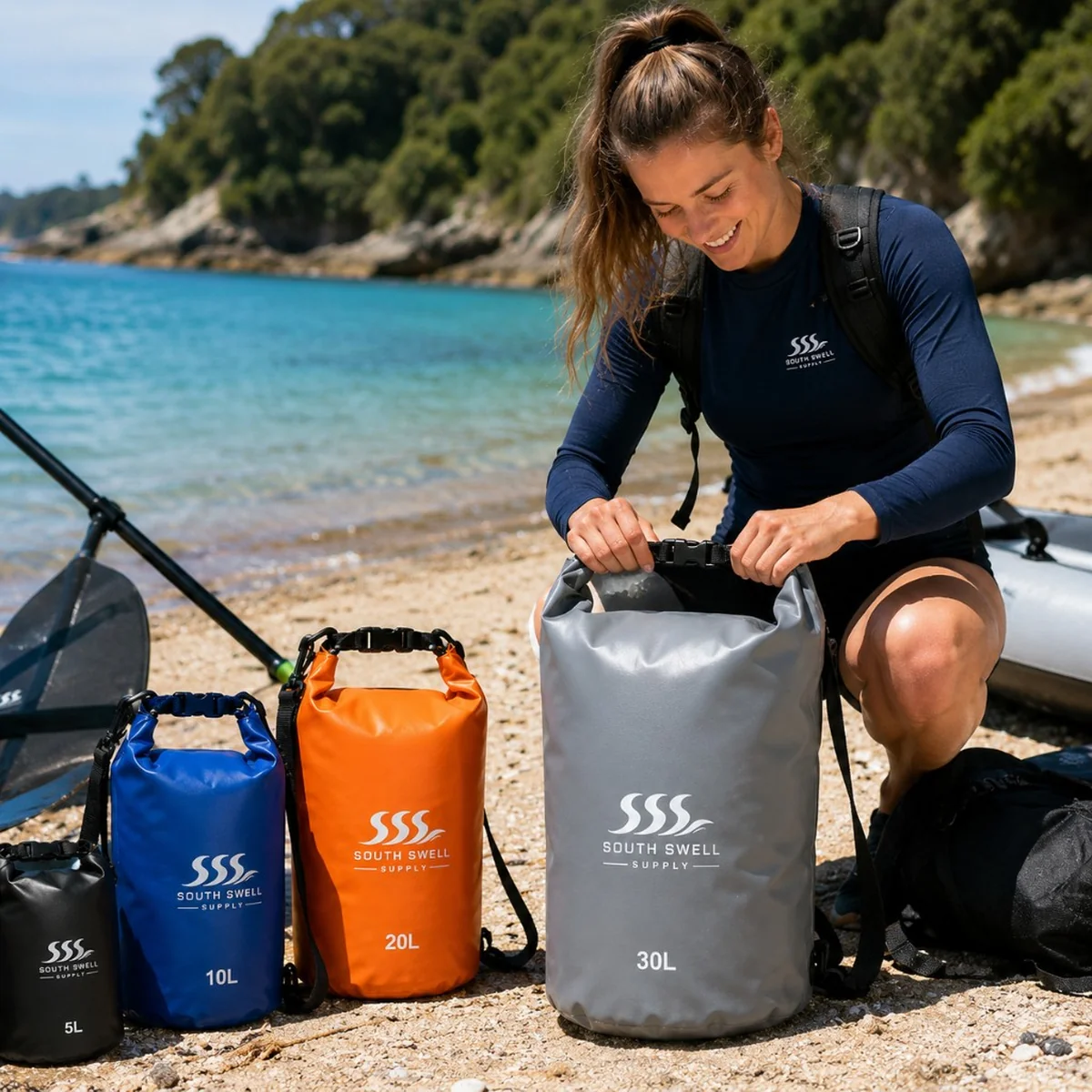 Woman on a sunny beach sealing a South Swell Supply dry bag beside a full size range — lifestyle use for kayaking and watersports retailers in New Zealand and Australia.