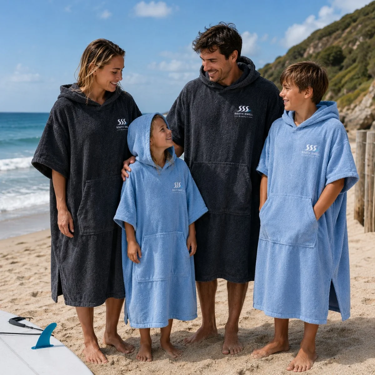 Family on a sunny beach wearing South Swell Supply hooded changing robes in charcoal and light blue — lifestyle use for surf schools and holiday parks in New Zealand and Australia.