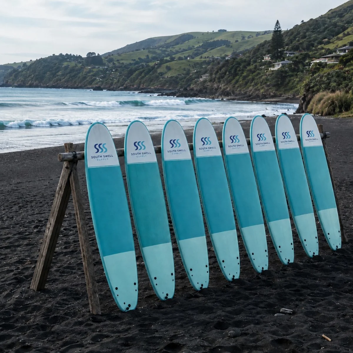 Fleet of eight custom-branded teal soft-top surfboards lined up on a black sand beach — bulk surf school and rental fleet orders for New Zealand and Australia.