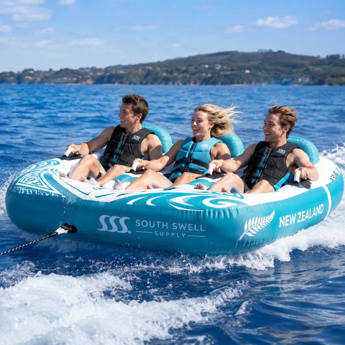 Three riders on a South Swell Supply 3-person towable tube being pulled across blue ocean water — lifestyle use for holiday parks, resorts and watersports rental fleets.